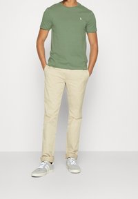 Vihreä puuvillainen t-paita, beiget chinot, harmaat tennarit. T-paidassa on pieni logo rinnassa. Rentoa, vartalonmyötäistä siluettia, hihat käännettyinä.