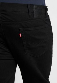 Los vaqueros de mezclilla negra presentan un parche de cuero en la cintura, una pequeña etiqueta de logotipo roja y un bolsillo trasero con costuras curvas. Tienen una textura suave y un corte recto.