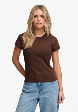Femme blonde portant un t-shirt marron moulant à manches courtes et un jean bleu clair, debout devant un fond blanc.