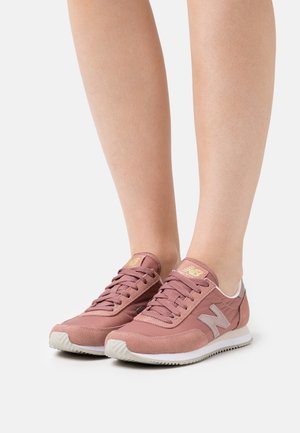 Fødder iført lyserøde New Balance-sneakers med hvide såler, stående på en ensartet lys baggrund.