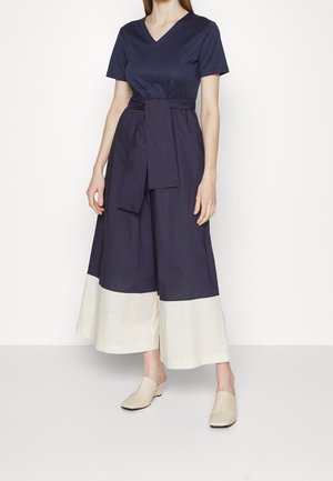 Kvinde iført en mørkeblå jumpsuit med brede ben og hvide manchetter, et taljebånd, korte ærmer og beige slip-on sko.