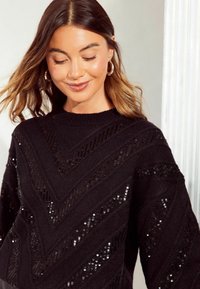 Maglione nero con un motivo a zig zag, caratterizzato da un lavoro a maglia testurizzata e dettagli in paillettes. Le caratteristiche includono un collo a giro e maniche morbide.