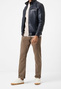 Man med marinblå läderjacka, beige tröja, bruna byxor och vita sneakers med gummisulor står mot en vit bakgrund.