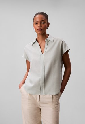 Kobieta o krótkich włosach, ubrana w jasnoszarą, krótkorękawową bluzkę z kołnierzykiem i beżowe spodnie, stojąca z rękami w kieszeniach na jednolitym tle.