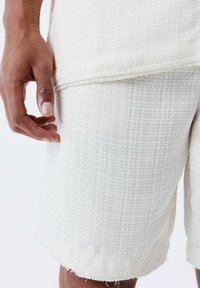 Bermudas blancas texturizadas con un patrón tejido, que presentan dobladillos deshilachados. La tela parece liviana y adecuada para el clima cálido.