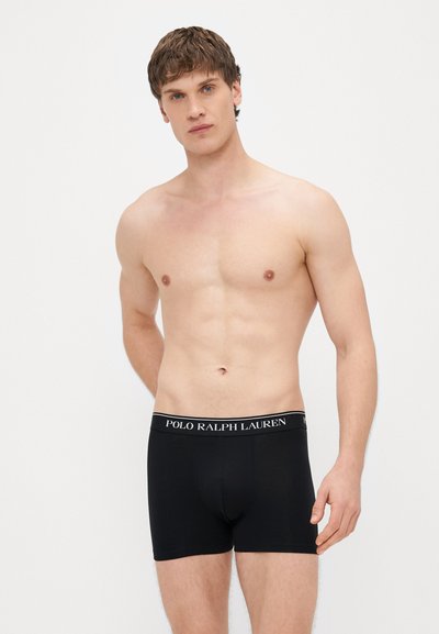 Čierne bavlnené boxerky s elastickým pásom so značkou „POLO RALPH LAUREN“. Model má krátke vlasy a stojí bez trička.