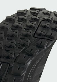 Semelle de chaussure de sport noire avec des crampons profonds et angulaires et des motifs texturés pour une meilleure adhérence. Bords renforcés et matériau flexible pour une durabilité accrue.