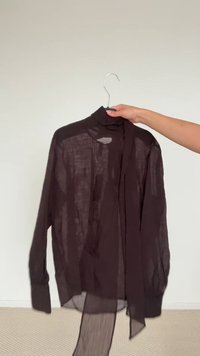Blusa nera trasparente a maniche lunghe con collo alto, caratterizzata da un laccio drappeggiato sul davanti e una vestibilità ampia, appesa a una gruccia metallica su uno sfondo neutro.