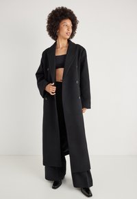Sort dobbeltrækket frakk med brede klapkraver, lange ærmer og lige snit, vist over en sort crop top og vide bukser.