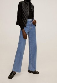 Svart vadderad väst över en svart polotröja, kombinerad med högmidjade ljusblå vida jeans och svarta loafers. Slät tyg med en avslappnad passform.