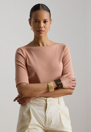 Femme aux cheveux plaqués en arrière portant un haut rose à encolure bateau, un pantalon crème taille haute, des boucles d'oreilles créoles dorées et deux bracelets, bras croisés.