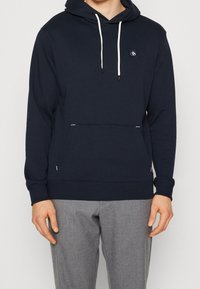 Sudadera azul marino hecha de una tela suave, con un bolsillo delantero, cordones y un pequeño parche de logo en el pecho. Los puños y el dobladillo son de canalé.