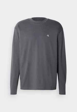 Siva majica dugih rukava napravljena od mekanog pamuka, s okruglim izrezom i izvezenim logotipom na prsima. Glatka tekstura s minimalnim detaljima šavova.