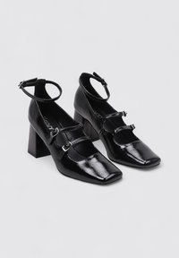 Zapatos de tacón bloque de charol negro con puntera cuadrada, que presentan tres correas y hebillas ajustables en los tobillos. Textura suave y acabado pulido.