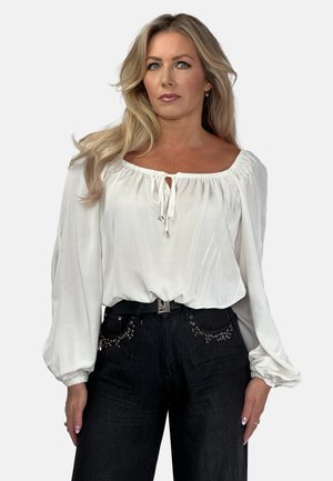 White, long-sleeve blouse with a wide neckline and tie detail at the front, paired with high-waisted black jeans featuring decorative studs.