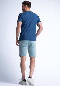 Mannelijke marineblauwe T-shirt met een textuurpatroon, gecombineerd met lichtblauwe opgerolde shorts en witte sneakers. Achteraanzicht toont informele kleding.