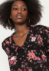 Blusa preta com estampado floral rosa; possui decote em V, mangas curtas e detalhes em botão na parte da frente. Textura de tecido suave.
