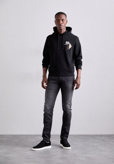 Svart hoodie med framficka, ribbade ärmslut och dragsko. Kombination med mörkgrå jeans och svarta sneakers med vita sulor.