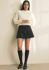 Elfenbensfarvet kabelstrikket cropped sweater med frynser, parret med en sort plisseret miniskørt og knæhøje sorte sokker. Sorte sko med spænde detaljer.