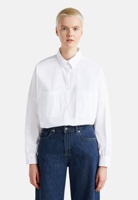 Camicia bianca cropped con bottoni, due tasche frontali, maniche lunghe e colletto classico, abbinata a jeans in denim blu a vita alta.