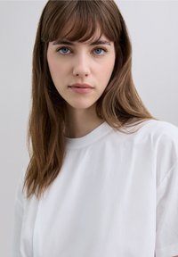 Weißes T-Shirt mit kurzen Ärmeln und rundem Ausschnitt, glatter Textur und leicht lockerem Sitz, getragen von einer Person mit langen, braunen Haaren.