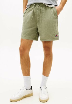 Uomo che indossa pantaloncini con coulisse verde oliva chiaro con logo Tommy Jeans, calzini bianchi corti e scarpe da ginnastica bianche, in piedi con le mani nelle tasche.