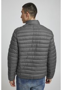 Grijze pufferjack met een horizontaal quiltpatroon, hoge kraag en een lichte glans. Gepaard met blauwe jeans, weergegeven vanuit de achterkant.
