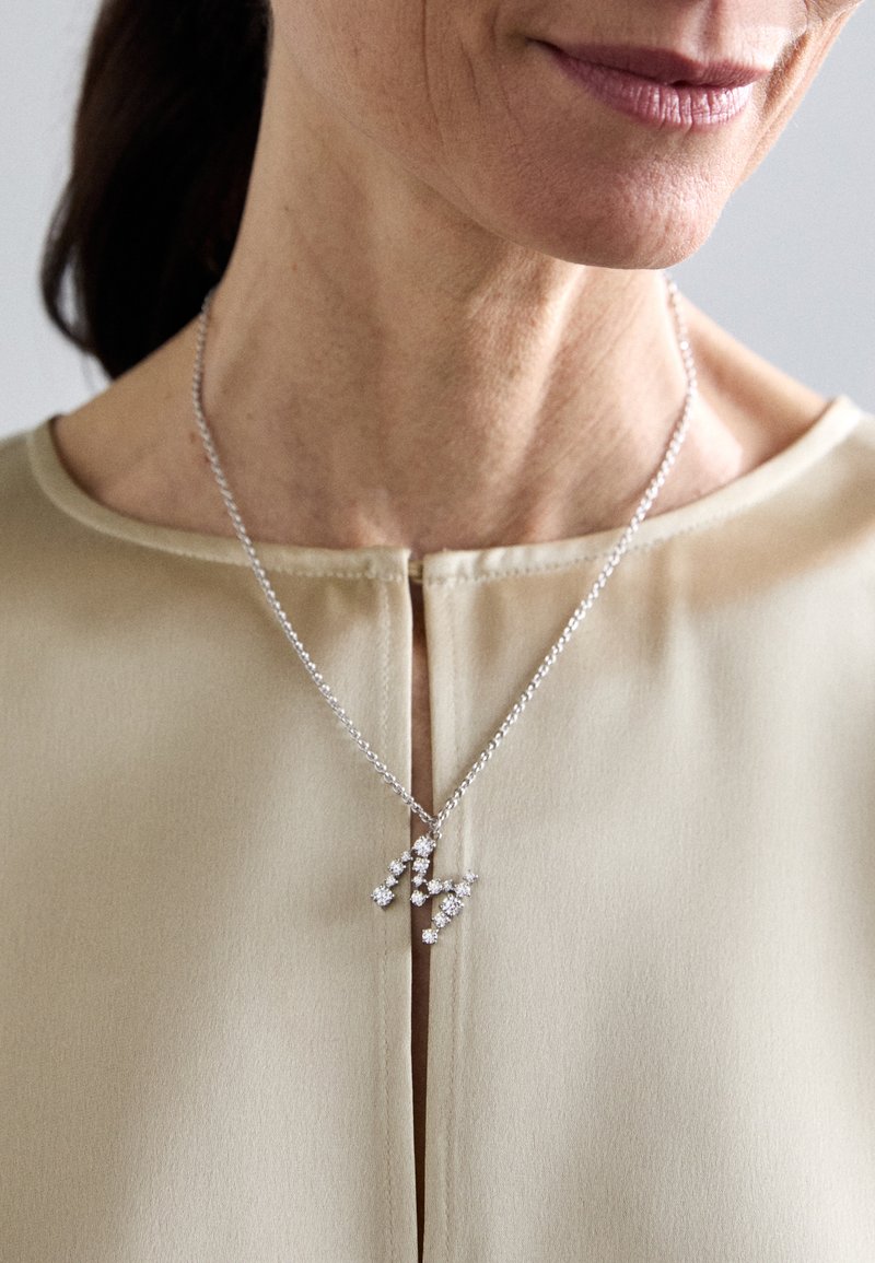 Ασημένιο κολιέ με κρεμαστά λουλουδάκια που διαθέτουν μικρές πολύτιμες πέτρες, φορεμένο πάνω από ένα ανοιχτό μπεζ μπλουζάκι με στρογγυλή λαιμόκοψη.