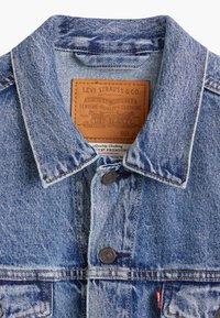 Jeansjacka i ljusblå färg, klassisk krage, knapplåsning, synliga sömmar och en läderetikett som anger märke och storlek M.