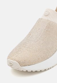 Slip-on sko i lys beige med teksturert stoff dekket av små, blanke perler. Har hvit såle og ribbet strikket ankel detaljer.