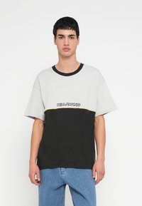 Γκρι και μαύρη μπλούζα με μπλοκ χρωμάτων και κίτρινη λεπτομέρεια· διαθέτει στρογγυλή λαιμόκοψη και άνετη εφαρμογή. Το λογότυπο της μάρκας εμφανίζεται στο μπροστινό μέρος.