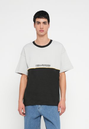 T-shirt em blocos de cinza e preto com detalhe amarelo; apresenta gola redonda e corte descontraído. Logótipo da marca exibido na parte frontal.