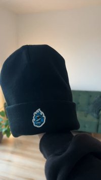 Căciulă neagră tricotată, cu o grafică cusută albastră și albă reprezentând gura unei creaturi și detalii de flăcări pe partea din față. Textură moale.