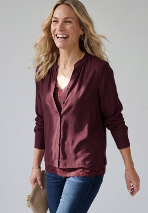 Lachende vrouw met lang blond haar, dragend een bordeaux blouse en kanten top, blauwe jeans, houdt een beige clutch vast tegen een effen achtergrond.