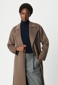 Cappotto in lana marrone con colletto a punte, maniche larghe e vestibilità rilassata, abbinato a un dolcevita a coste blu navy e pantaloni grigi.