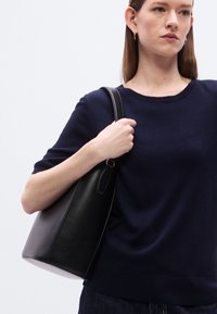 Μαύρη τσάντα tote από δέρμα με λεία υφή και στρογγυλεμένο σχήμα, με μία κορυφαία λαβή και ελάχιστα μεταλλικά στοιχεία.