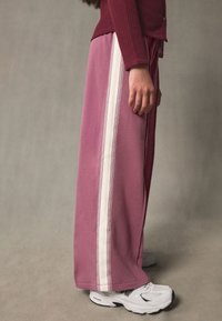 Lyserøde, løstsiddende joggingbukser med en hvid lodret stribe langs siden. Bruges med hvide træningssko, der viser en afslappet pasform og tekstur.