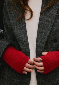 Personne portant des gants rouges en tricot sans doigts, tenant les revers d'un blazer gris texturé sur un haut blanc.