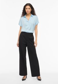 Ανοιχτό μπλε πουκάμισο με κοντά μανίκια και κουμπιά, σε συνδυασμό με μαύρο παντελόνι ψηλόμεσο, φαρδιάς γραμμής. Μαύρες γόβες με χρυσές λεπτομέρειες στο εξάρτημα.