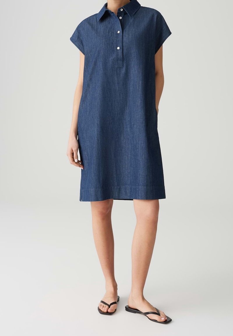 Femme portant une robe chemise en denim bleu foncé sans manches avec col et des sandales plates noires, debout devant un fond clair uni.