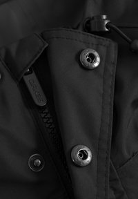 Primer plano de una chaqueta de tela negra que muestra una cremallera con el nombre de la marca y botones de presión metálicos con patrones de estrellas.