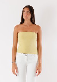 Top giallo senza spalline con una finitura strutturata e luccicante. Abbinato a jeans bianchi, indossato da una persona in piedi contro uno sfondo semplice.