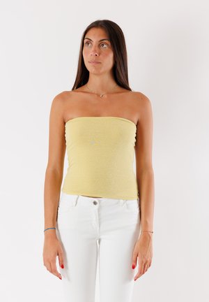 Top giallo senza spalline con una finitura strutturata e luccicante. Abbinato a jeans bianchi, indossato da una persona in piedi contro uno sfondo semplice.