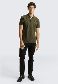 Olivgrünes Polohemd mit weißem Kragen, kurzen Ärmeln und einem Logo auf der Brust, kombiniert mit schwarzen Slim-Fit-Jeans und schwarzen Schuhen.