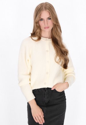 Crème-kleurige geribbelde cardigan met een ronde hals, vier knopen aan de voorkant en opgerolde mouwen, gecombineerd met een zwarte rok met hoge taille.