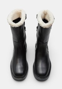 Botas de tobillo de cuero negro con un mullido puño de piel de oveja blanca, cierre lateral con cremallera y un acabado liso y mate. Suelas de goma resistentes.
