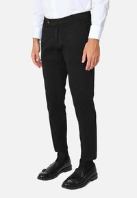 Pantaloni neri in cotone con una vestibilità slim, tasche frontali e chiusura con bottone. Indossati con scarpe nere e calzini neri con motivo.