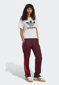 Weißes Kurzarm-T-Shirt mit schwarzem Adidas-Logo, kombiniert mit burgunderfarbenen Hosen mit roten Seitenstreifen und weißen Sneakers mit schwarzen Streifen.
