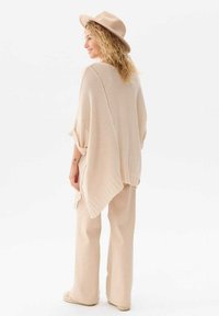 Beżowy dzianinowy poncho z otwartymi rękawami i prążkowanym dołem, zestawione z pasującymi szerokimi spodniami i miękkim kapeluszem. Widoczne subtelne detale tekstury.