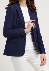 Blazer marine en tissu texturé, avec fermeture à un bouton, revers crantés et poches à rabat, porté sur une chemise blanche.